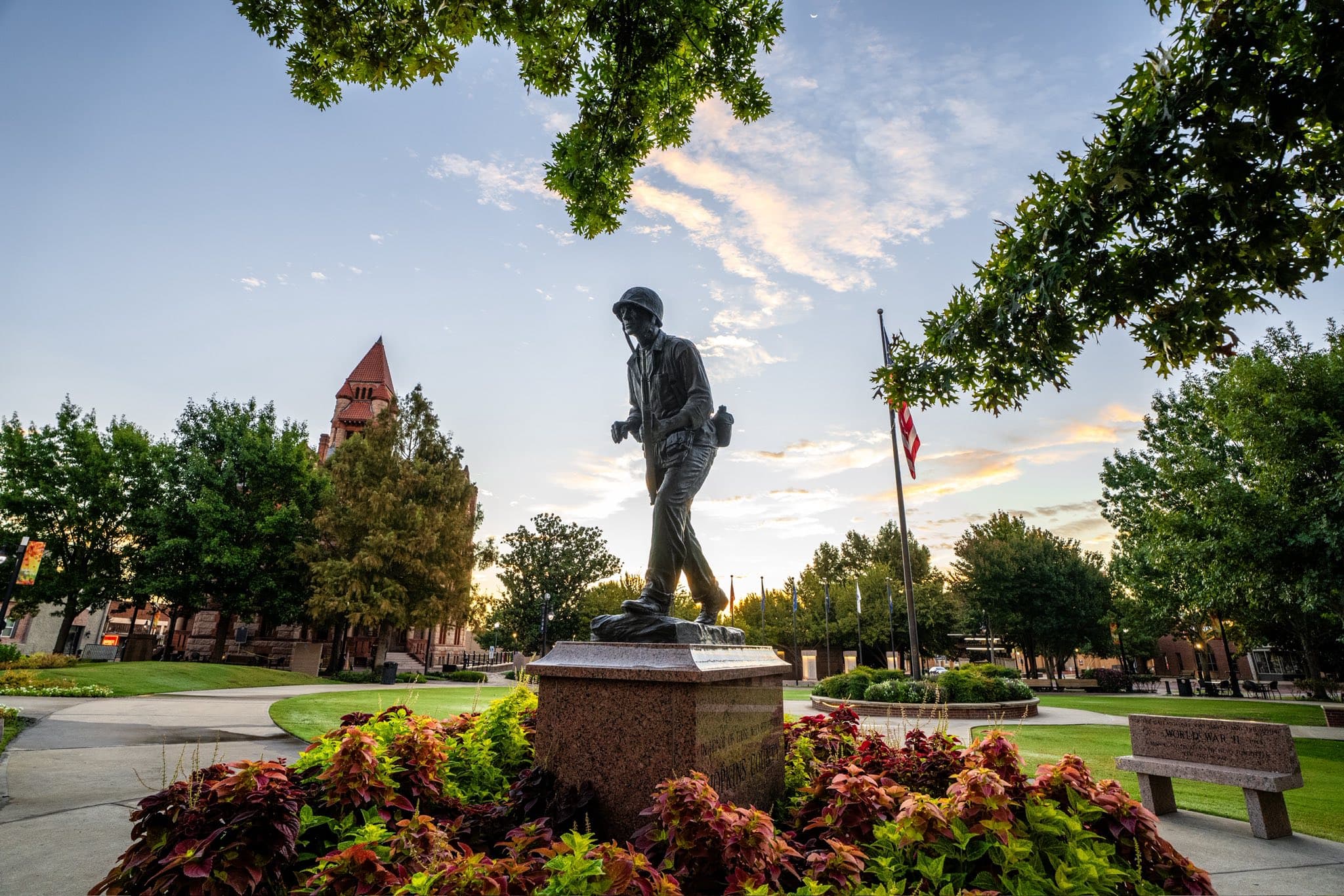 A soldier statue stands in a park among greenery and a colorful flower bed, with a building and flags in the background under a twilight sky.